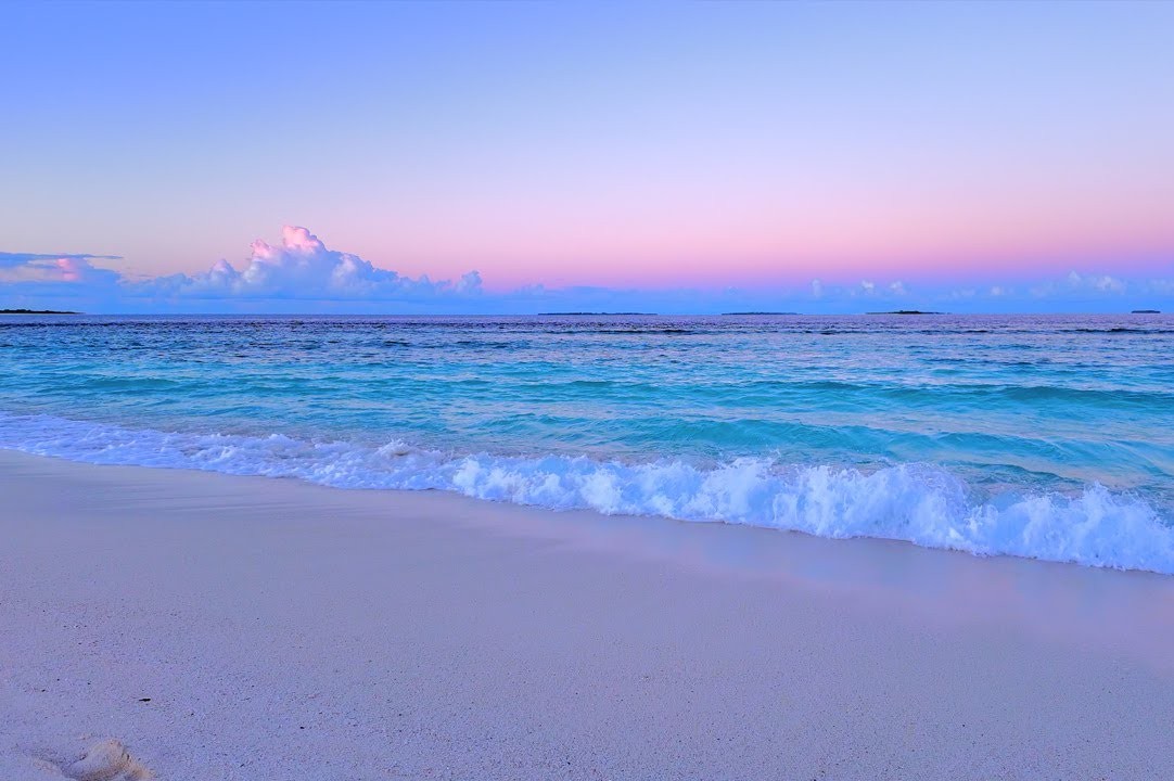 Empty sandy beach at sunrise with turquoise waves, pink sky reflections on wet sand, and distant land on the horizon